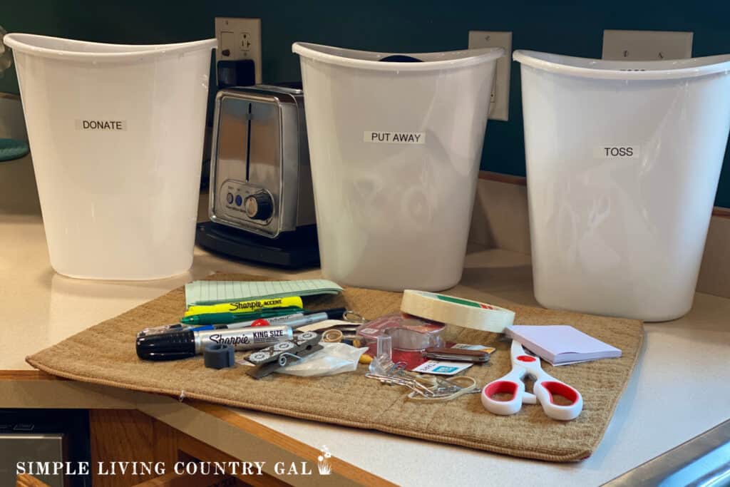 3 labeled buckets in front of a pile of clutter on a kitchen counter