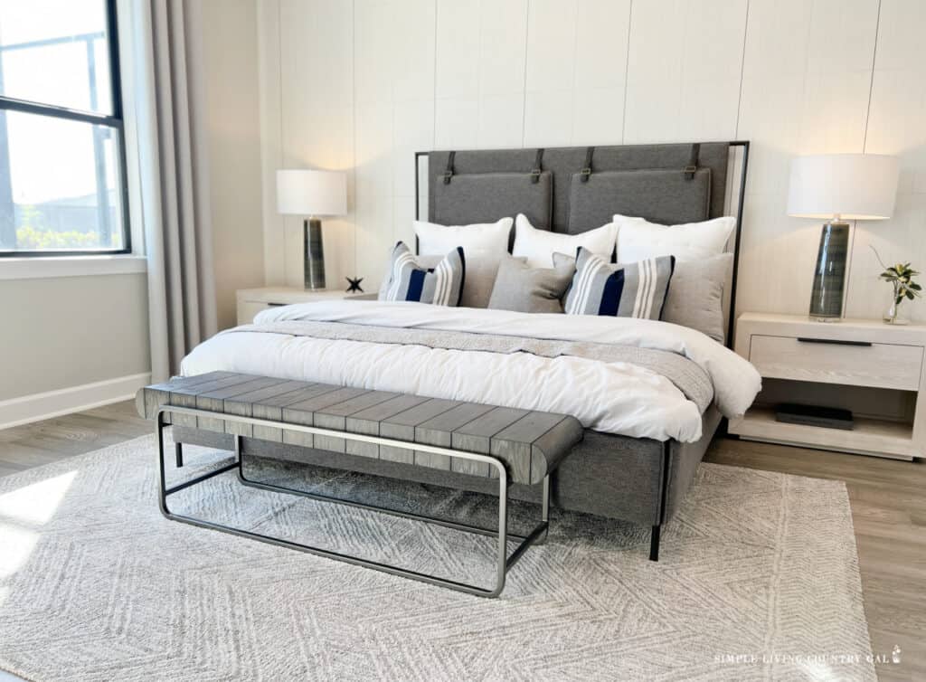 A grey bed with a foot stool and a white rug