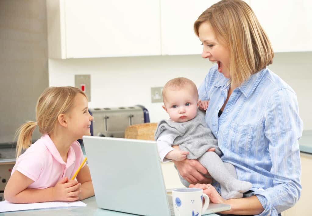 woman holding a toddler laughing with a little girl with a laptop in a kitchen
