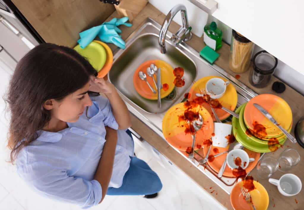 woman with hand on head looking at sink full of dirty dishes