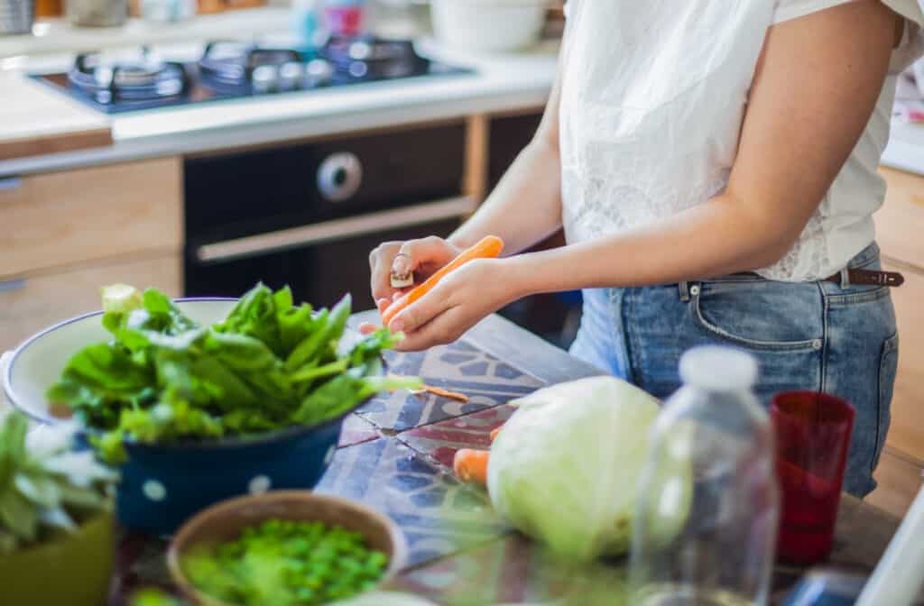 woman peeling a carrot with other vegetables on a cutting board around her. 