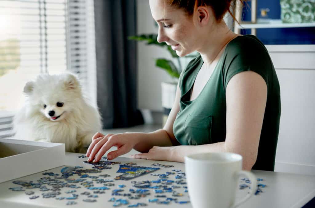 woman next to a little white dog working on a puzzle