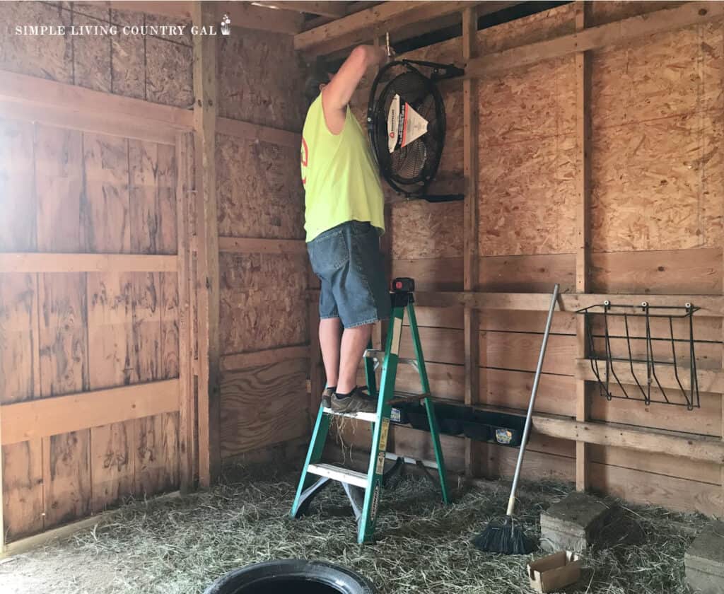 a man on a ladder hanging a fan in a barn