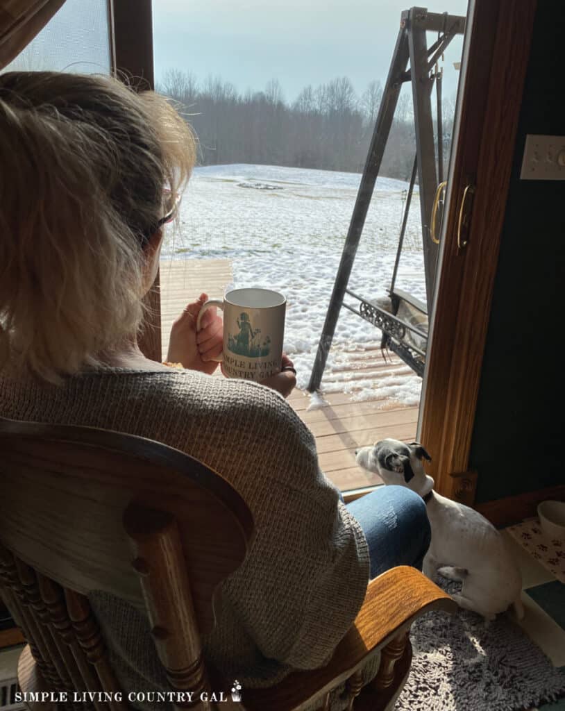 a woman drinking coffee looking out a window near to her dog