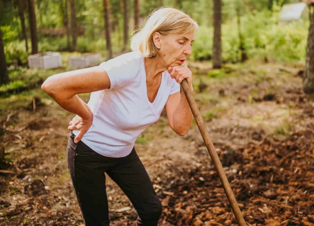 A woman in a white shirt leaning on a shovel holding her sore back