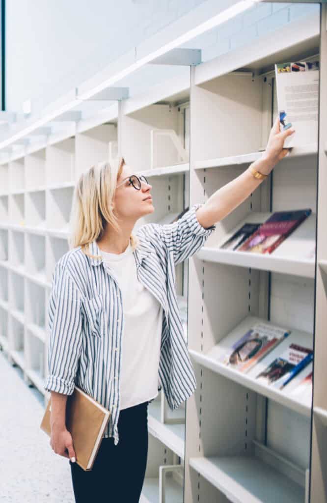 Young woman in casual clothes and glasses standing near bookcase and selecting book to read in a library