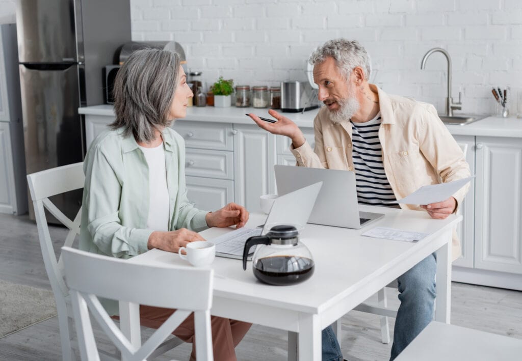 a midlife couple with laptops talking at a kitchen table