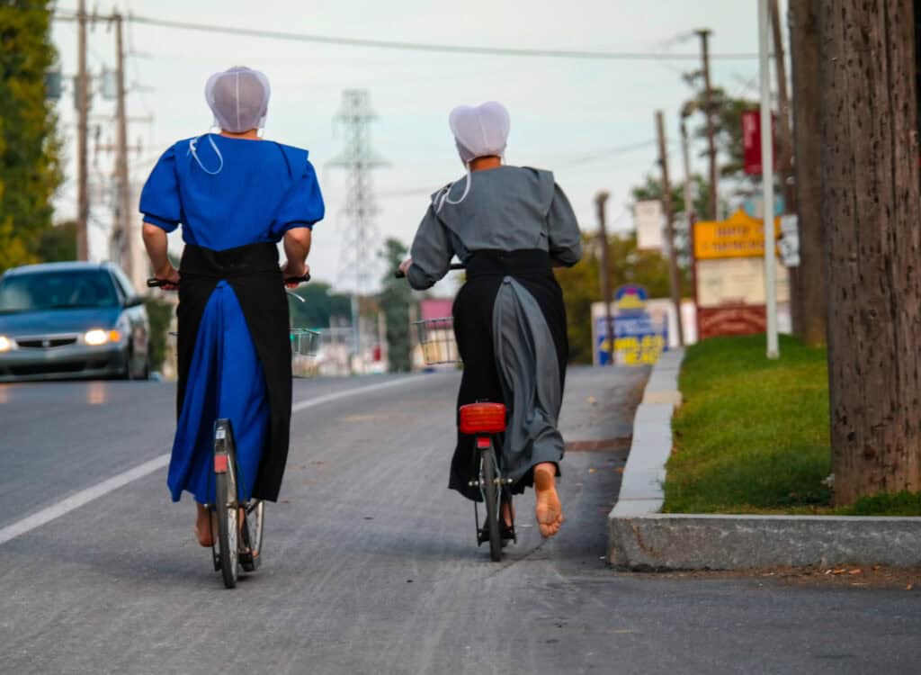 two amish girls riding bikes barefoot on a small town road 