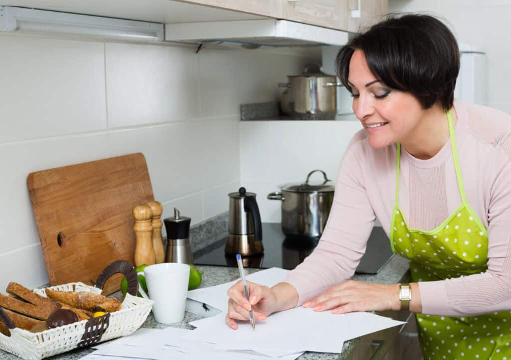 A woman in a green apron writing on papers in a kitchen, with a cutting board, spices, and a basket of bread on the counter